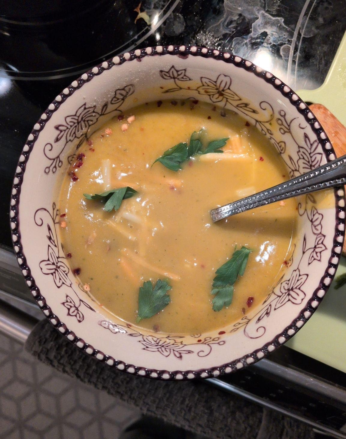 A decorated bowl contains a creamy yellow-orange squash soup garnished with parsley and red pepper flakes, with a spoon resting inside.