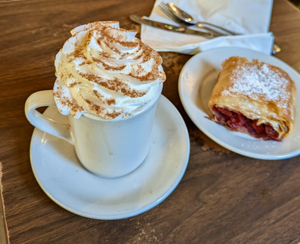 A mug of whipped cream-topped beverage is accompanied by a plate of pastry dusted with powdered sugar on a wooden table.