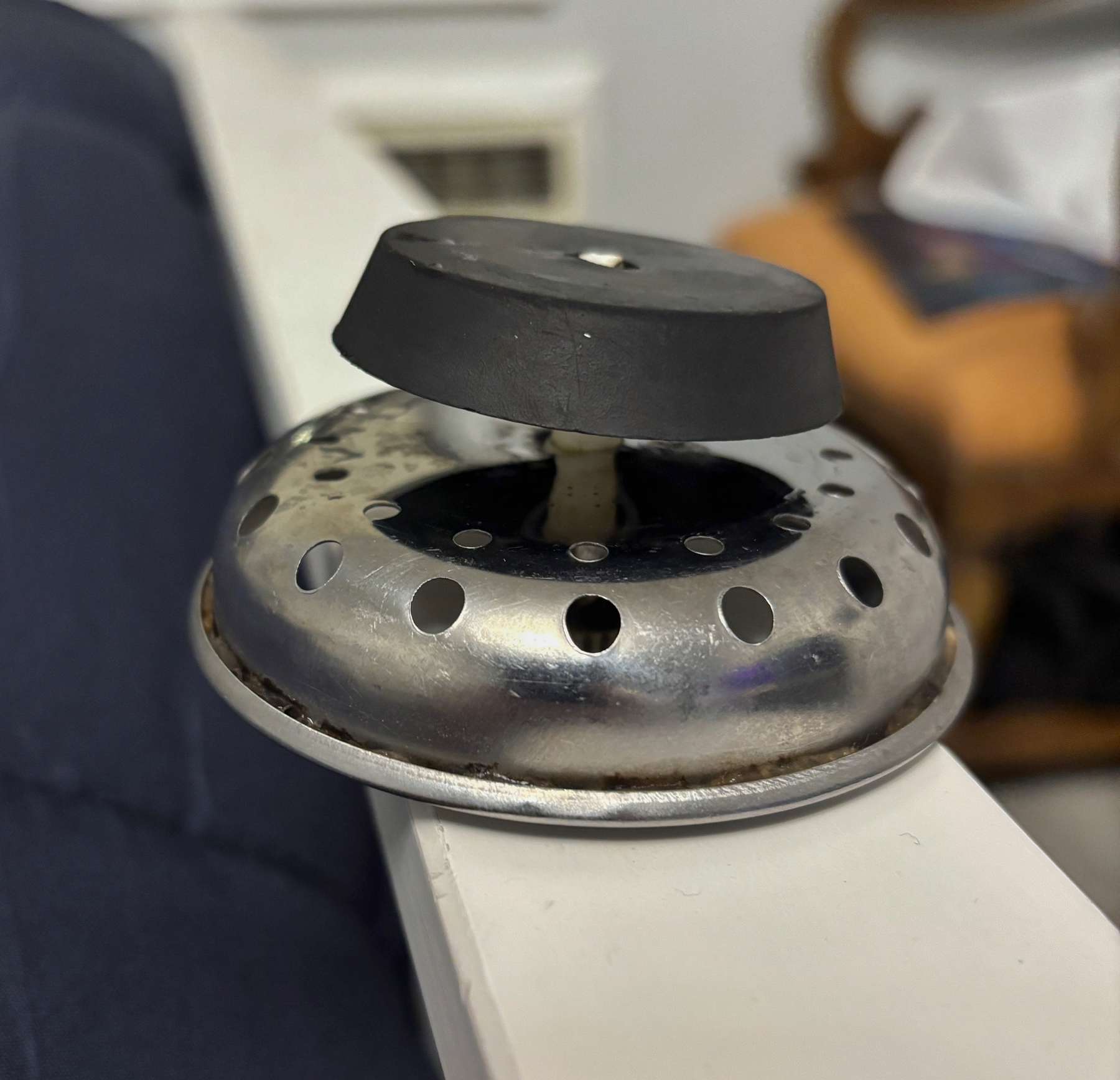A metal kitchen sink drainer is placed on a white surface upside down with a black rubber stopper attached to the top of it.
