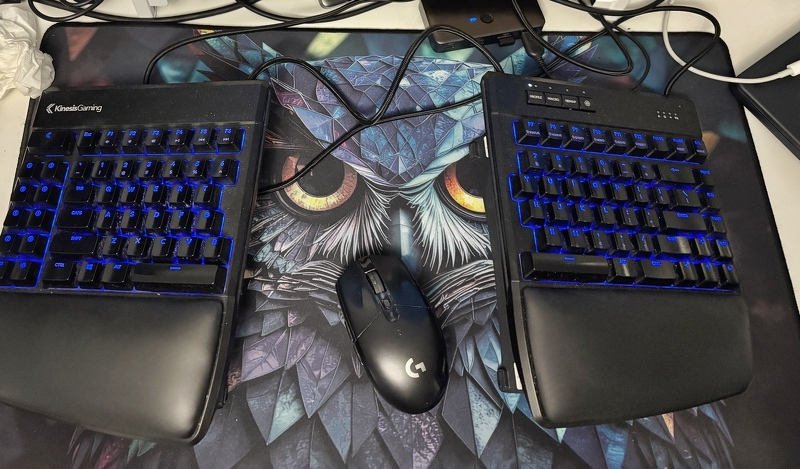 Auto-generated description: A split ergonomic keyboard with blue backlighting is connected by a cable, positioned on a large owl-themed mousepad alongside a Logitech gaming mouse.