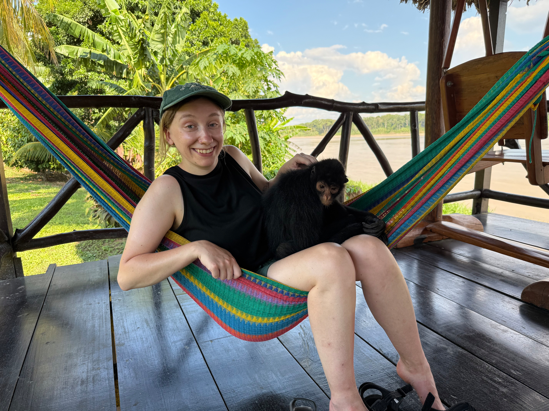 A person relaxing in a colorful hammock on a wooden deck, holding a small monkey, with lush greenery in the background.