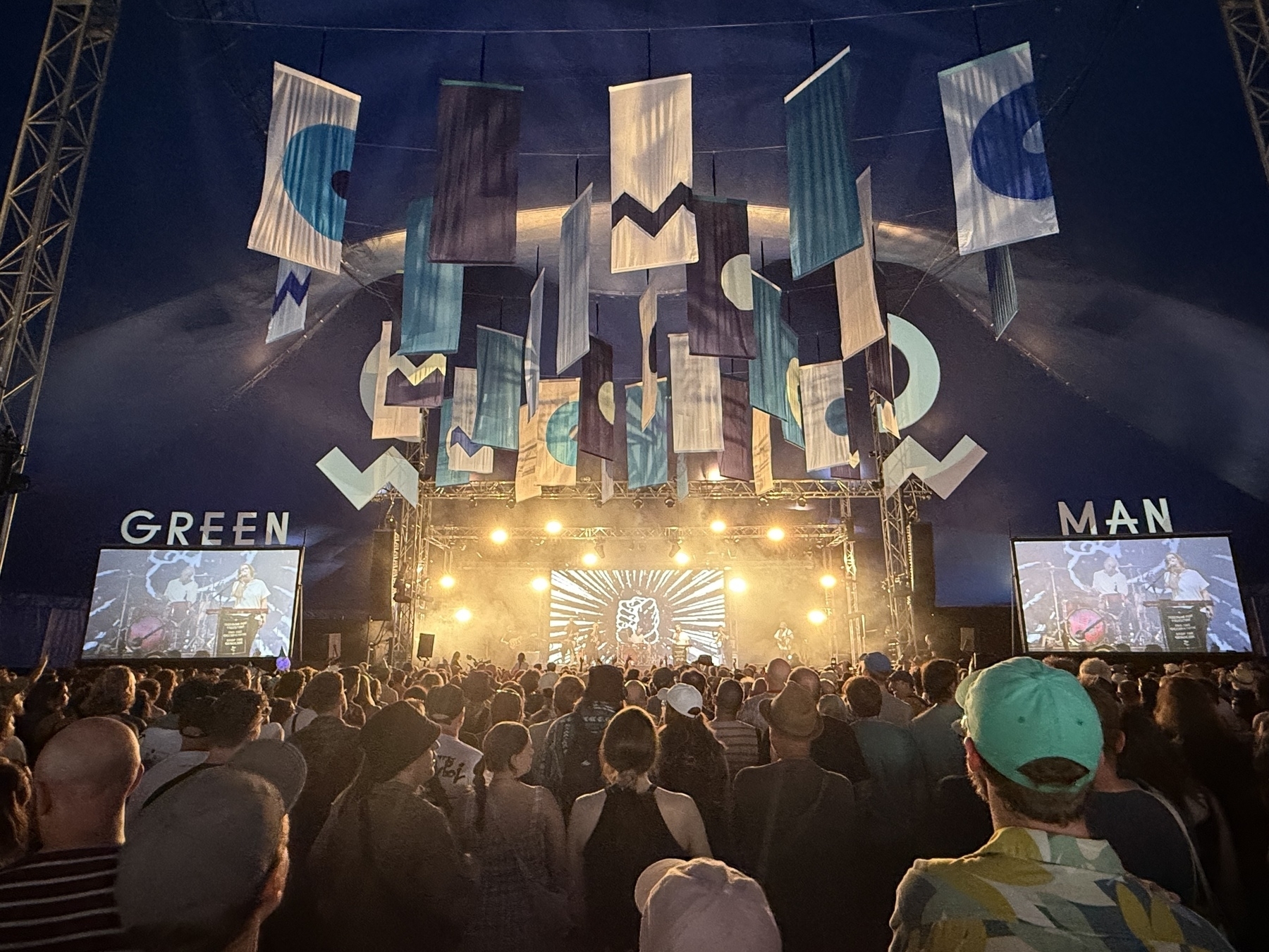 A crowd watches a performance on stage inside a large event tent with colorful banners and the words GREEN MAN displayed.