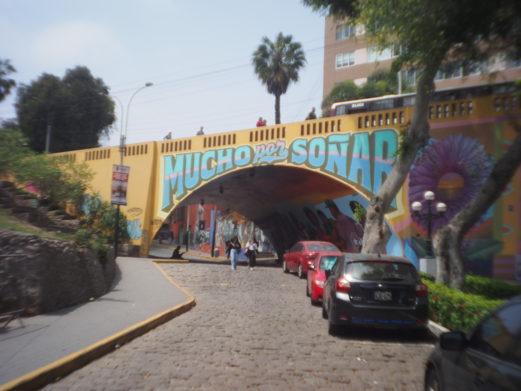 A colorful mural with the phrase MUCHO POR SOÑAR adorns a bridge over a cobblestone street with parked cars and trees nearby.