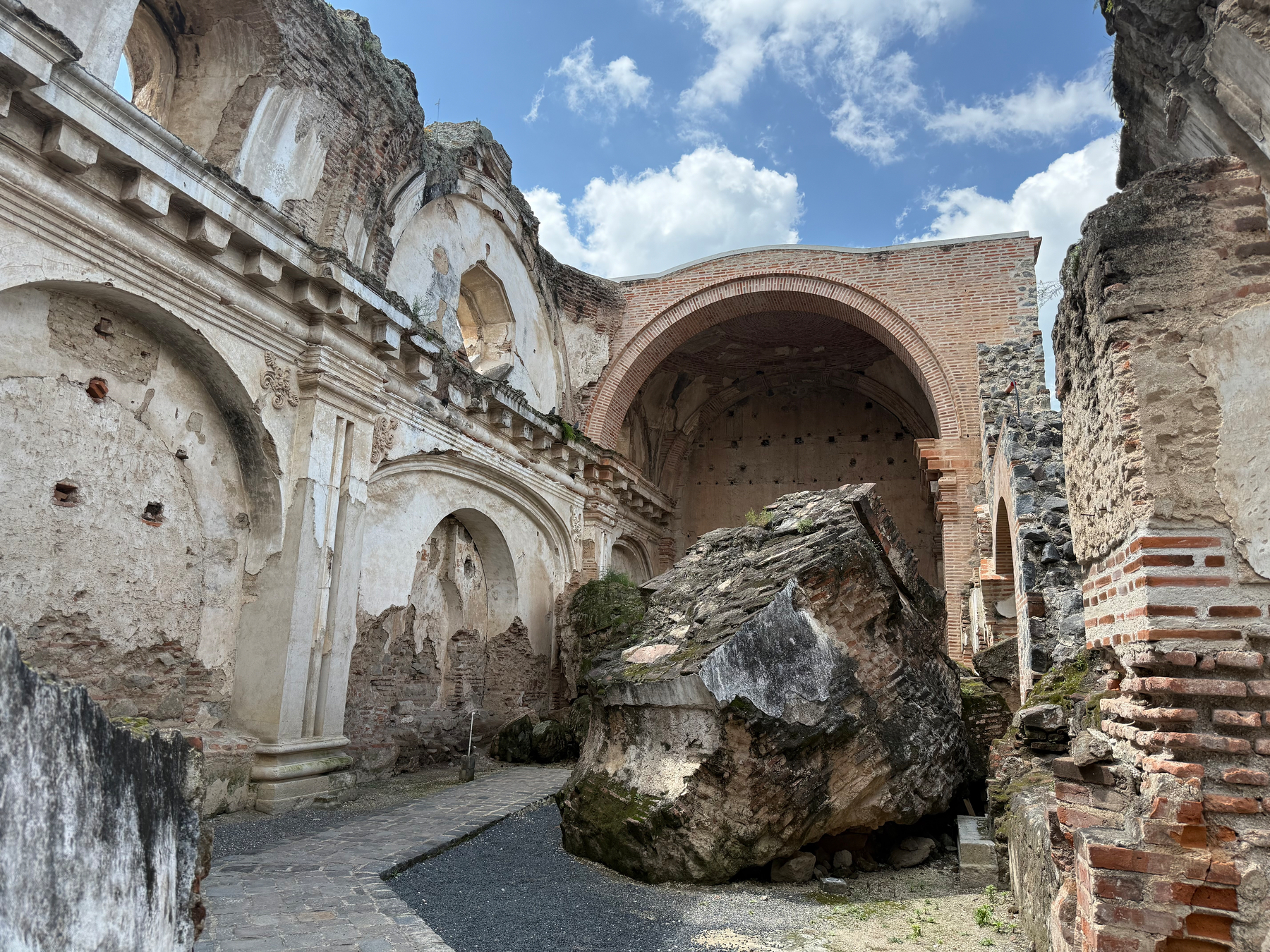 Auto-generated description: Ruins of an ancient stone structure feature partially collapsed walls, an archway, and a large fallen boulder under a blue sky with clouds.