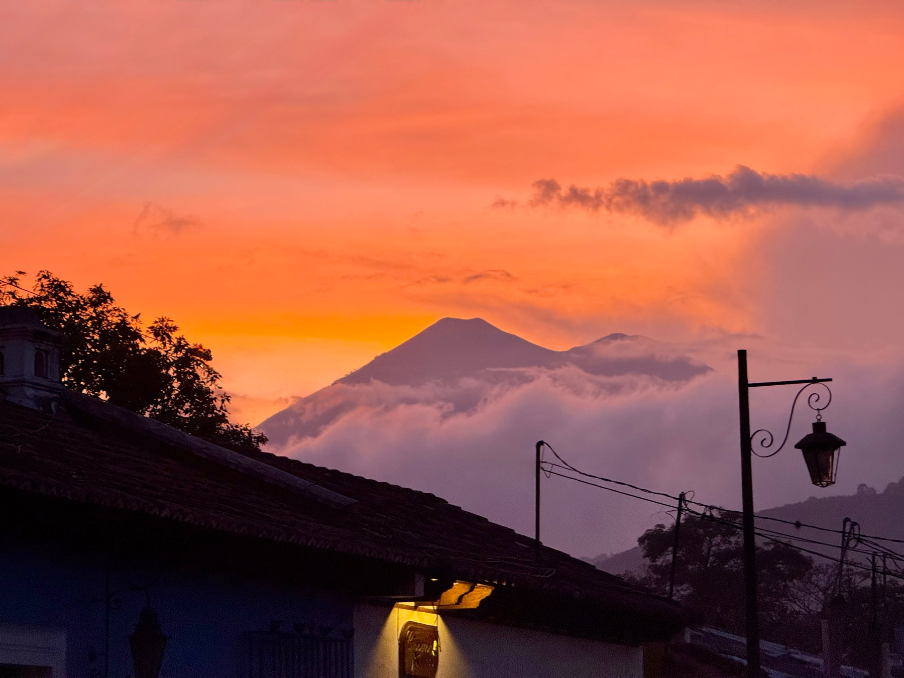 Auto-generated description: A picturesque sunset with vibrant orange and pink hues illuminates a silhouetted mountain and a charming village scene, complete with traditional rooflines and streetlights.