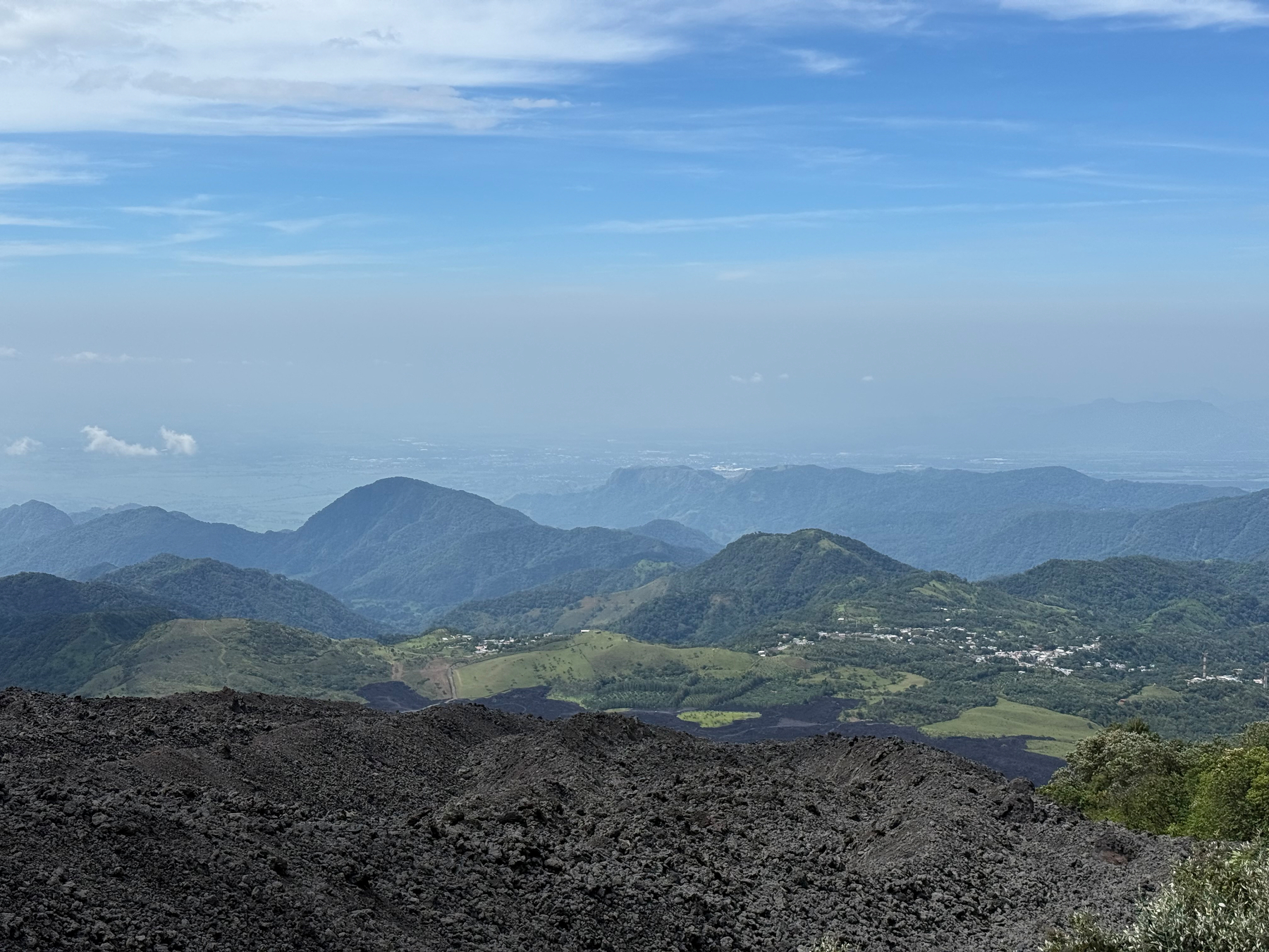 Auto-generated description: A landscape view showcases rolling green hills and mountains under a blue sky, with rough dark terrain in the foreground.