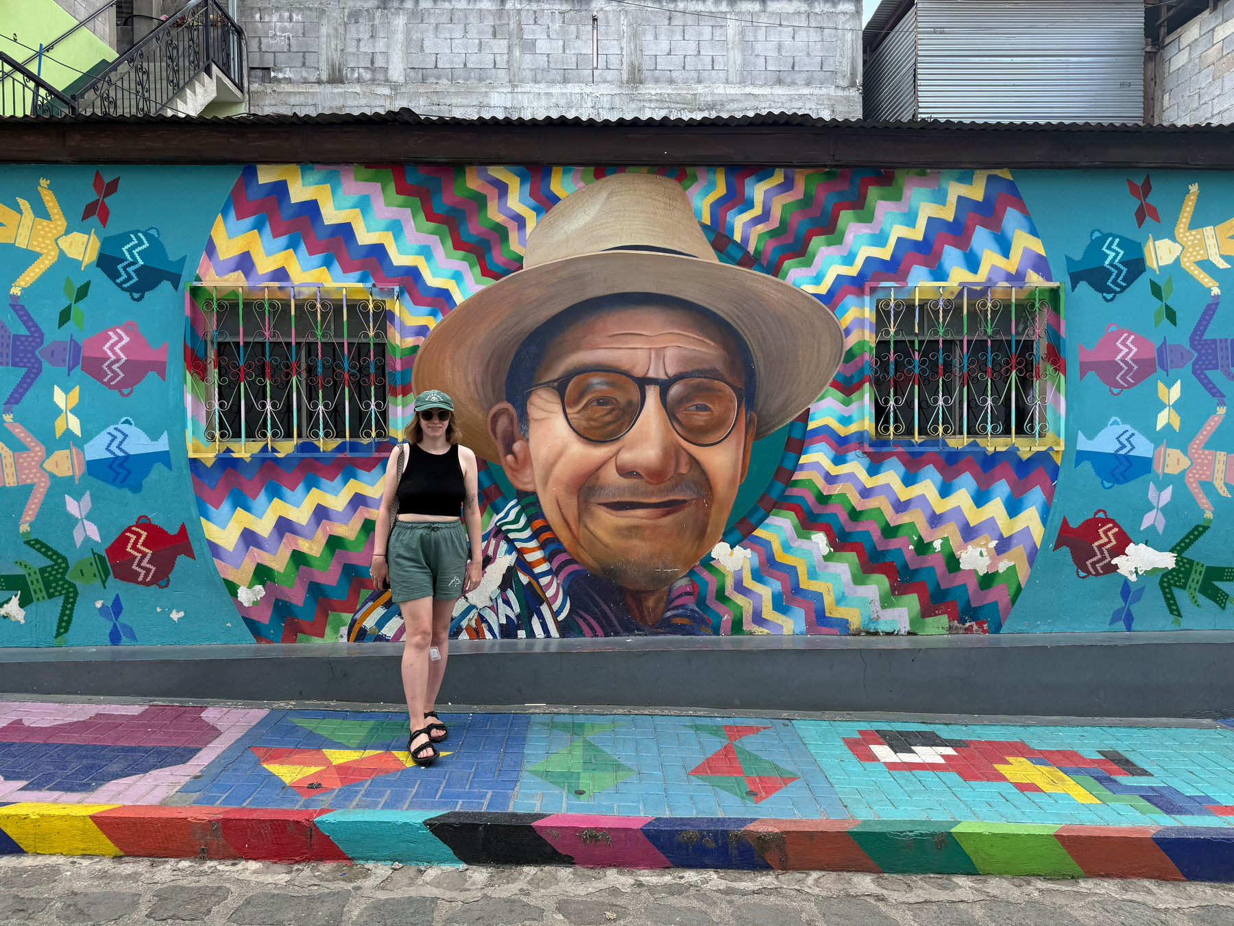 Auto-generated description: A person stands in front of a colorful mural featuring an older man wearing glasses and a hat.
