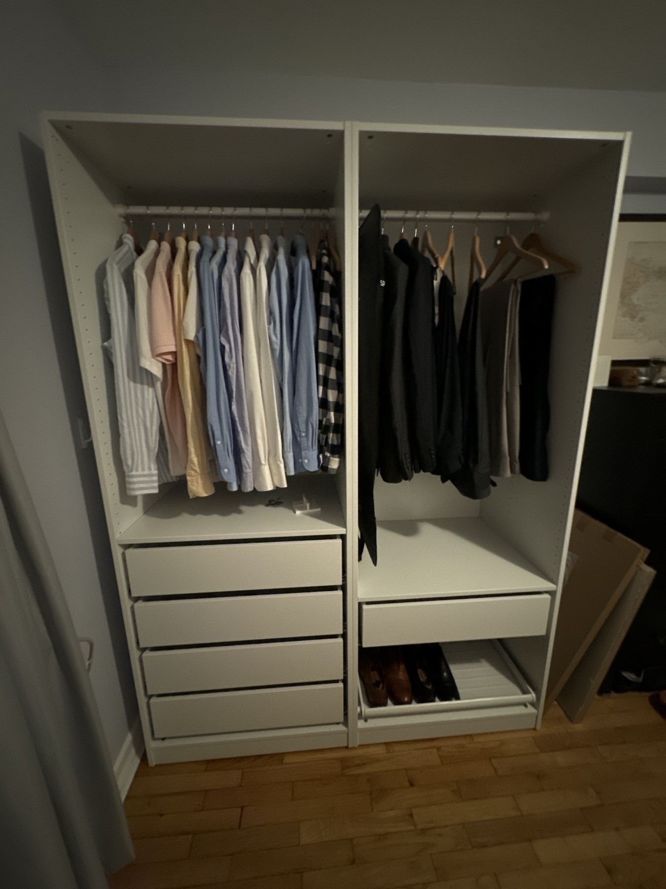 Image of an open IKEA Pax wardrobe. The left section displays an array of neatly hung shirts in pastel shades of pink, blue, and white. The right section contains darker clothing, including charcoal and navy suits and pants. Below the hanging clothes on the left is a set of white drawers, on the right is a shelf with two pairs of shoes neatly arranged.
