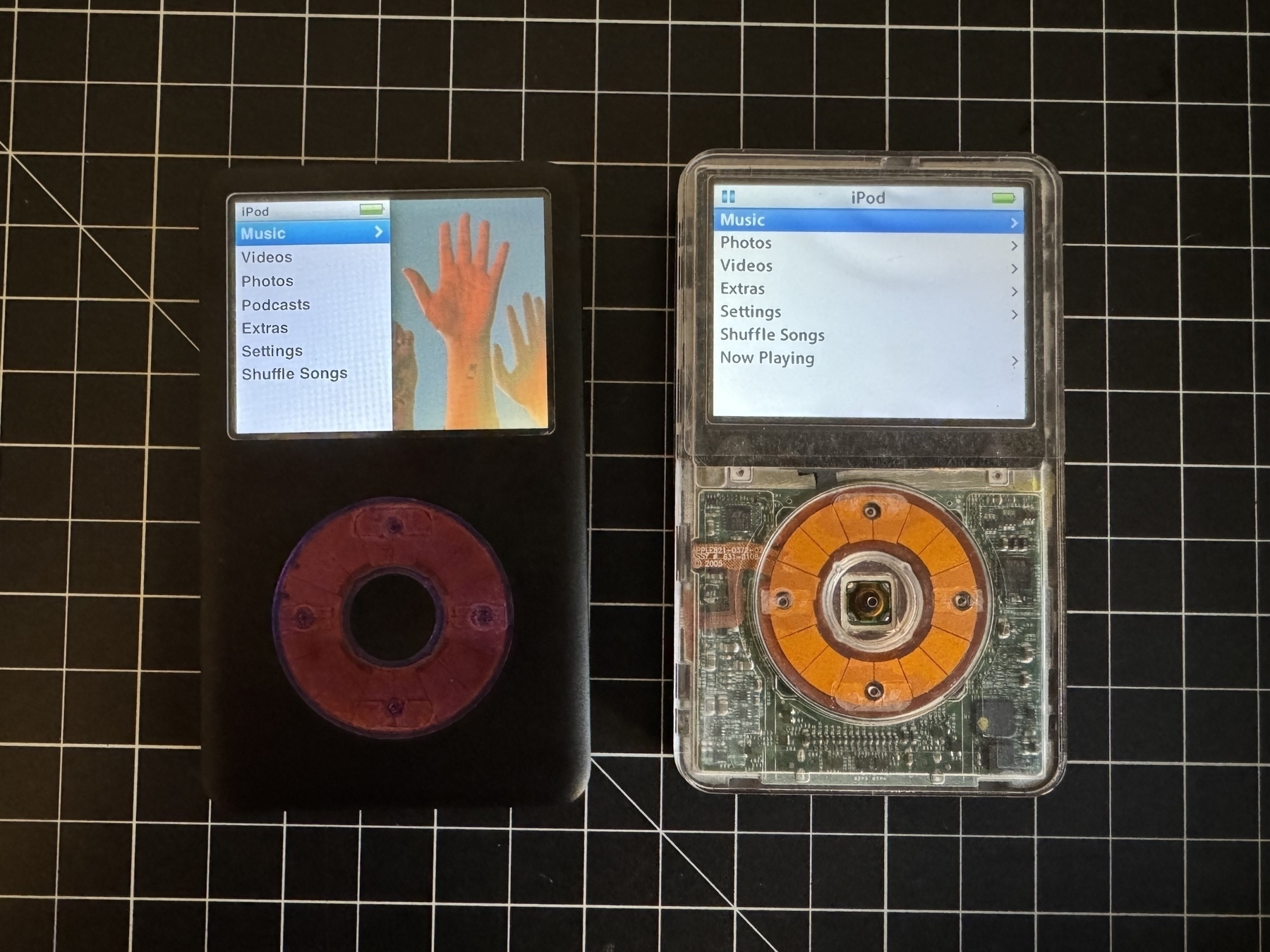 Two classic iPods next to each other on a workbench. The left one has a black aftermarket shell and purple click wheel, the right one has a clear shell and click wheel. 