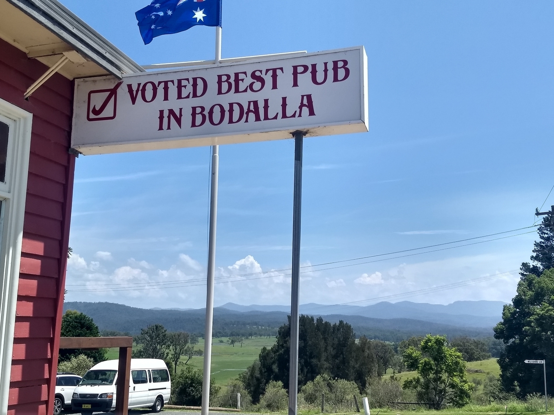 A view of green paddocks and distant forested hills from the verandah of a country pub on the South coast of NSW. A sign reads: Voted best pub in Bodalla. The Australian flag flies above.