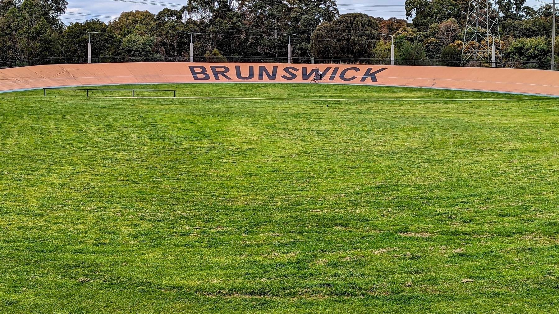 An outdoor velodrome in Melbourne, with an expanse of green grass in the foreground. A training cyclist is almost camouflaged by the large sign on the track: BRUNSWICK.