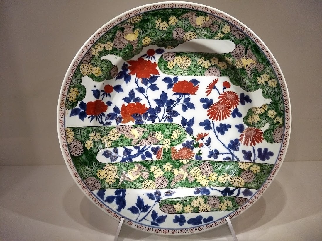 A large antique Japanese plate on gallery display. The design includes flowers, birds and clouds.