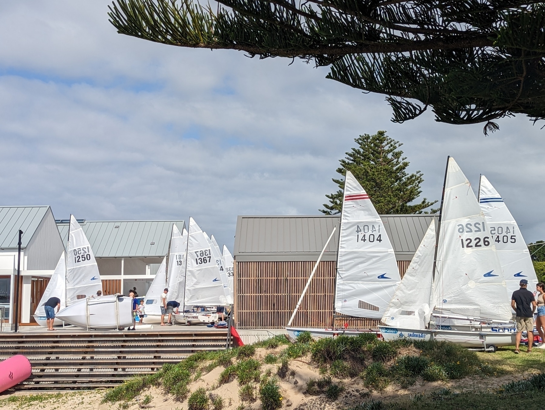 A flotilla of Flying Eleven sailing dinghies, white sails raised, cluster around a sailing club