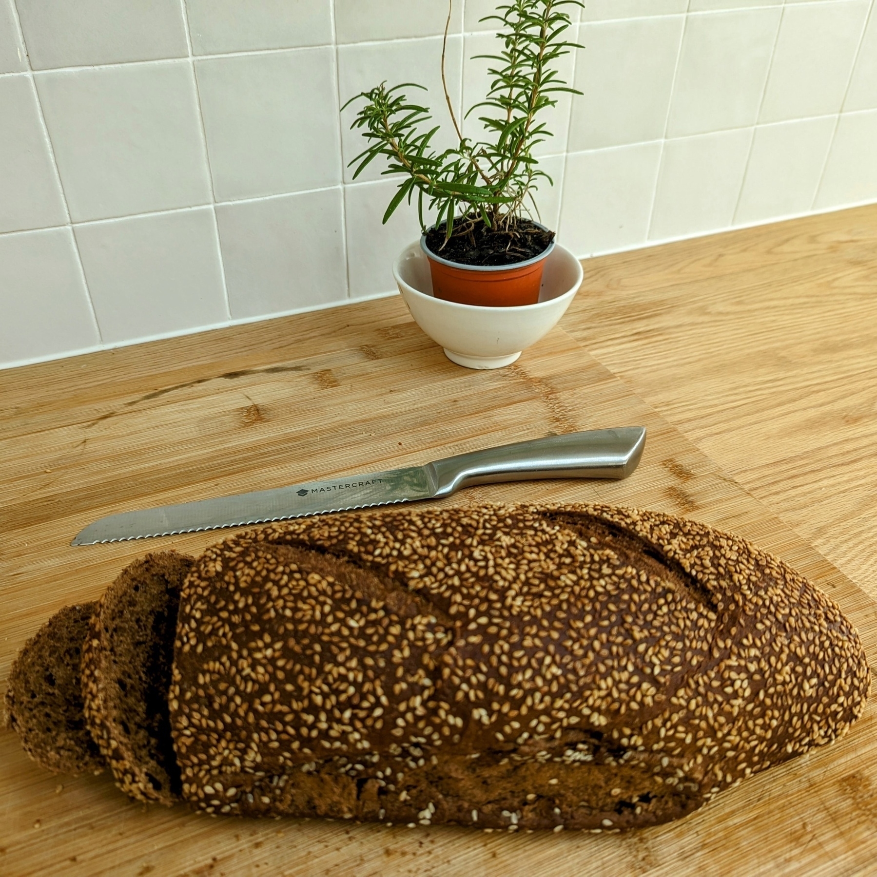 A wholemeal loaf on a bread board with a small potted rosemary plant behind. The loaf has a couple of slices already made.