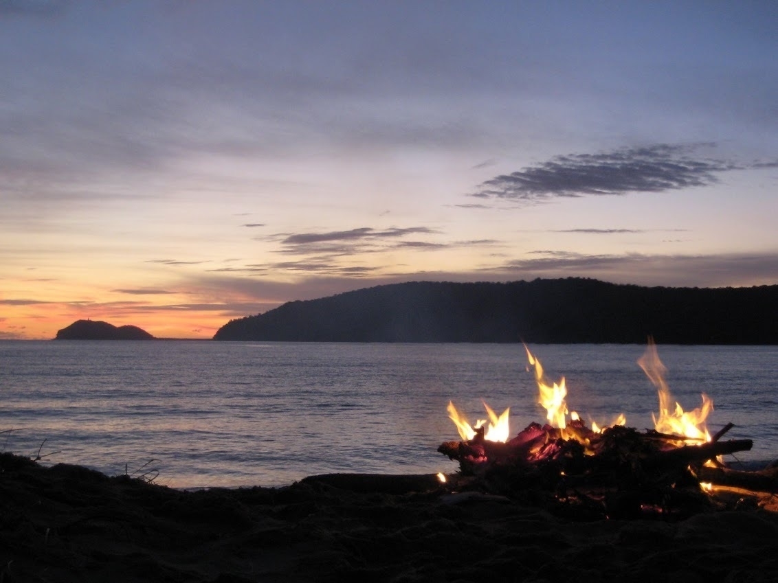 A campfire blazes on the shore at dawn. The sun is about to rise above a distant headland.