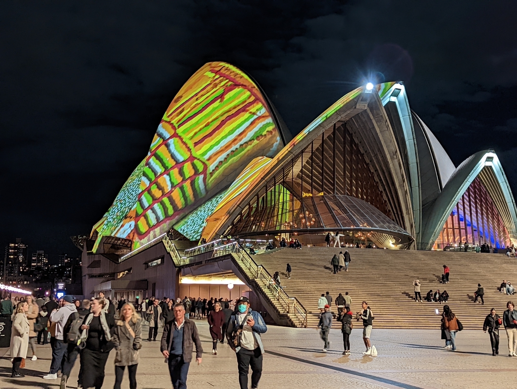 People walk past the front steps of Sydney Opera House. It’s dark and the  famous sails of the building’s roof are illuminated with an Aboriginal art motif.