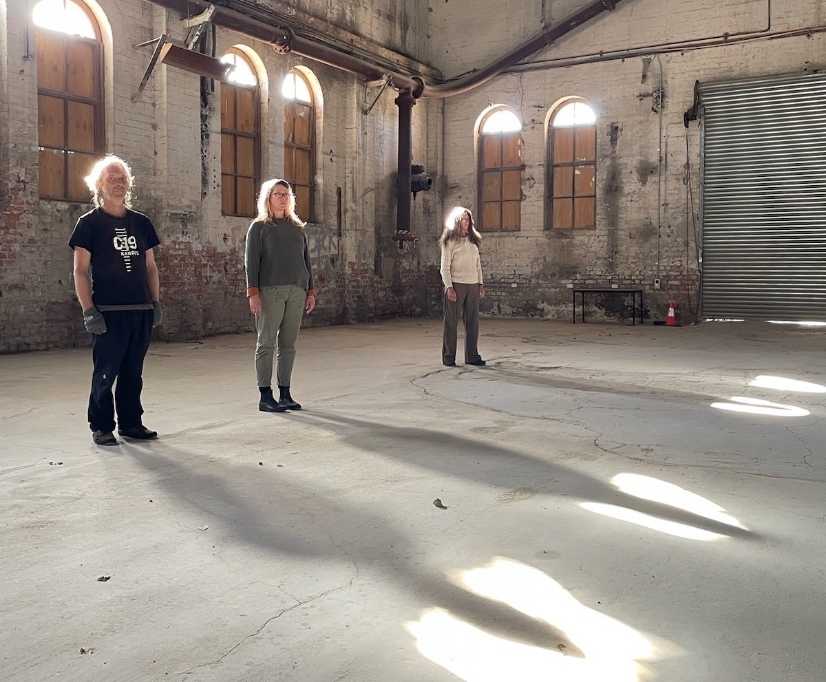 Three people stand in a line in the empty power house of the Foundations, a former cement works. Sunlight through arched windows creates a halo effect around their heads.