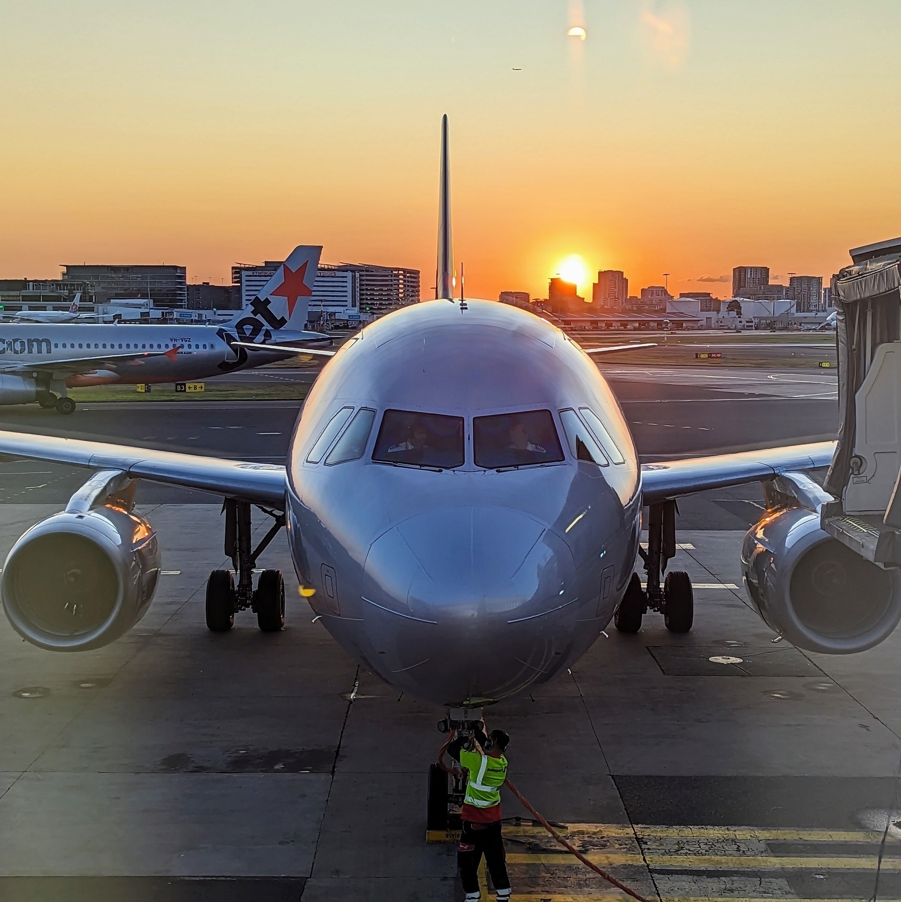 A photograph of the nose of a jet plane parked at an airport gate. In the background, the sun sets in an orange sky behind city apartment blocks.