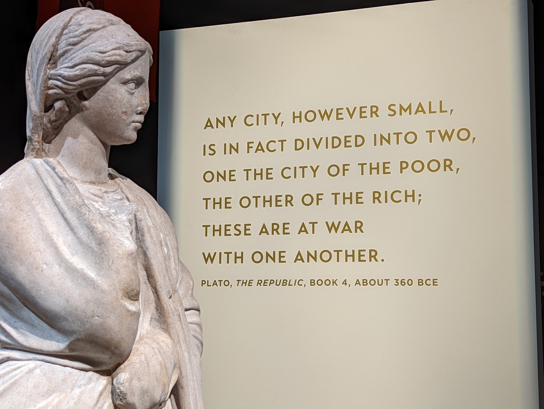 An ancient Greek marble statue of a woman regards a museum sign with a quote from Plato's Republic, which reads: Any city, however small, is in fact divided into two, one the city of the poor, the other of the rich; these are at war with one another.