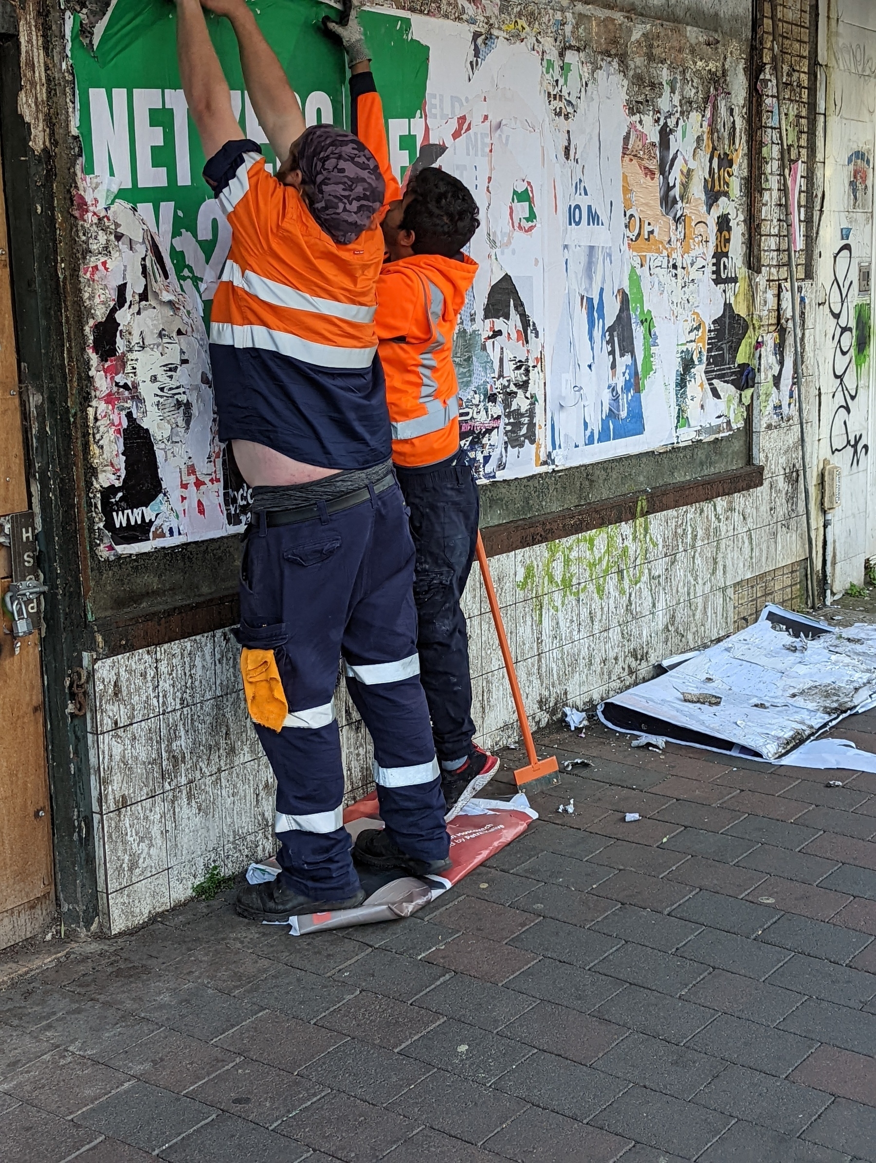 Two workers are pulling down old event posters off a billboard.