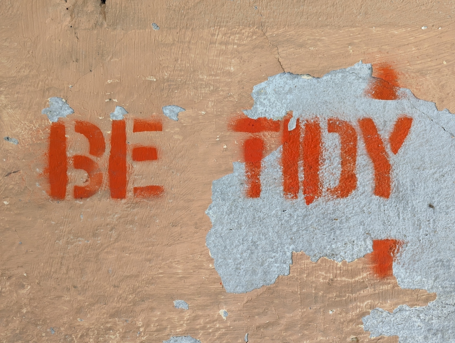 A stenciled sign painted in orange over old, peeling paint on a wall reads: Be tidy.