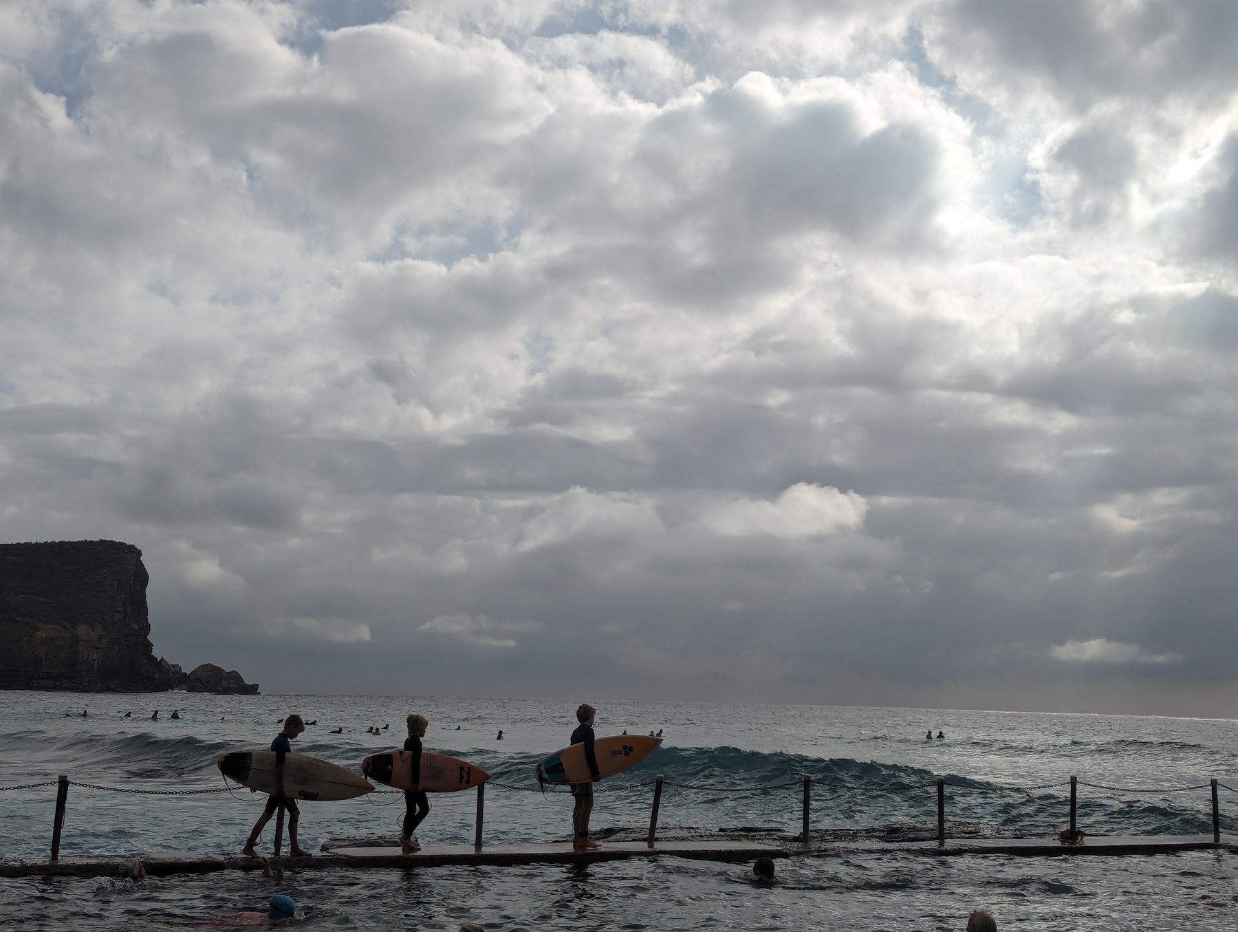 Three kids walking in a row with surf boards, with a stormy sky behind them.