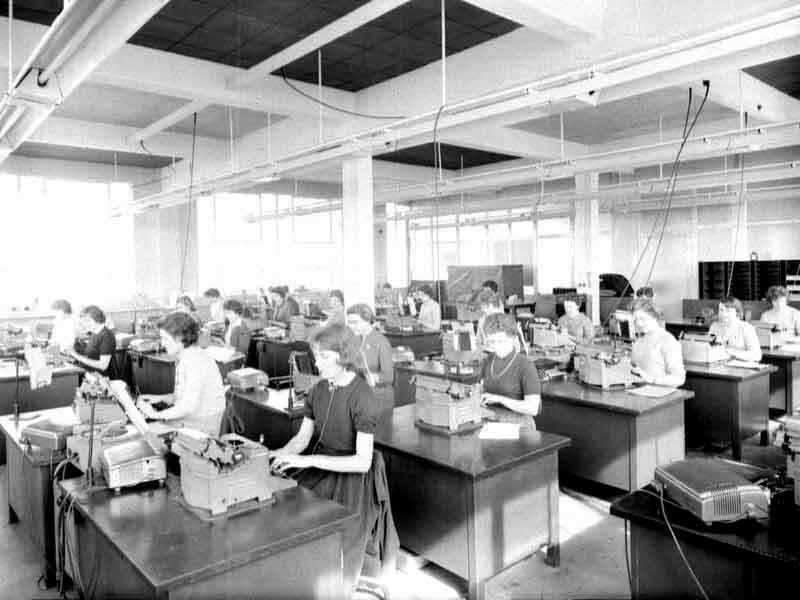 A black and white photo of a typing pool in the 1950s. Rows of women sit at rows of desks in an office with a typewriter each.