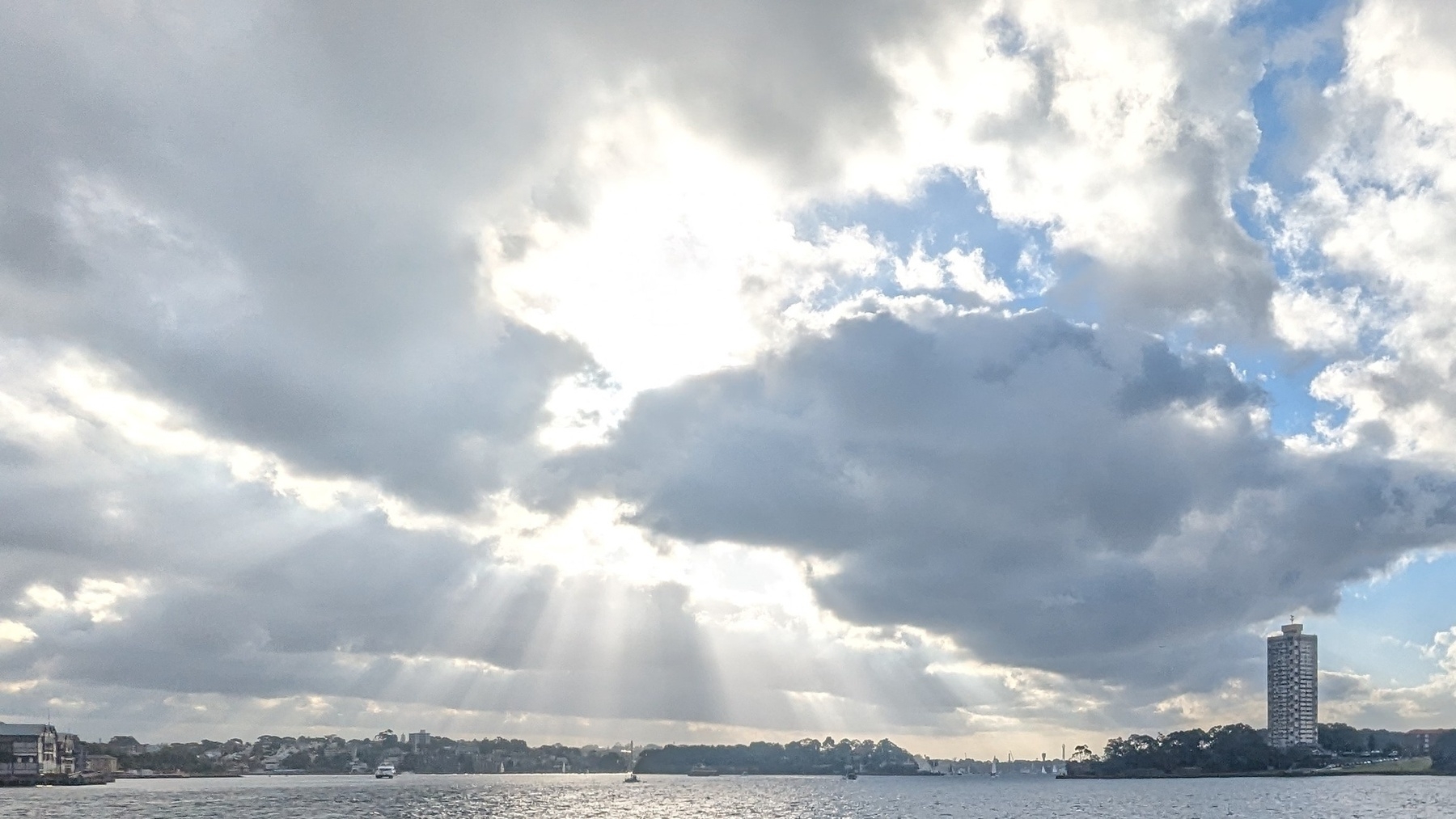 Rays of sunlight break through clouds over water, with Sydney's Blues Point Tower to the right in the middle distance 