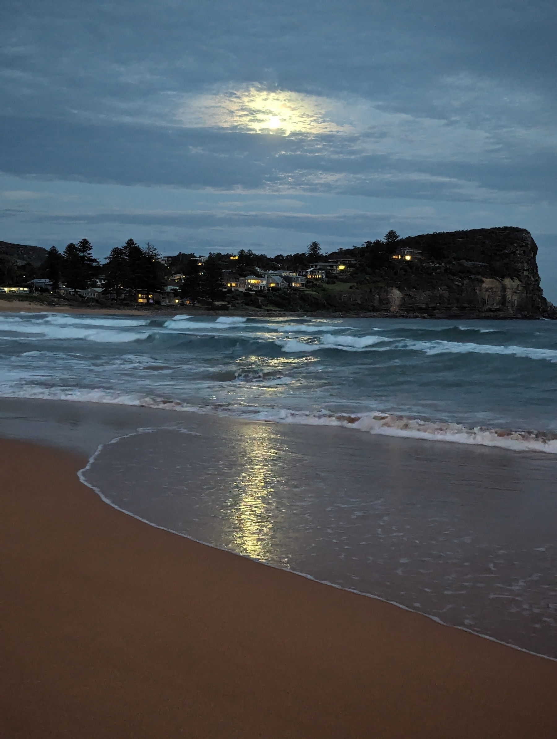 Moonlight through clouds above a rocky headland. In the foreground the moonlight is reflected in the waves lapping the shore.