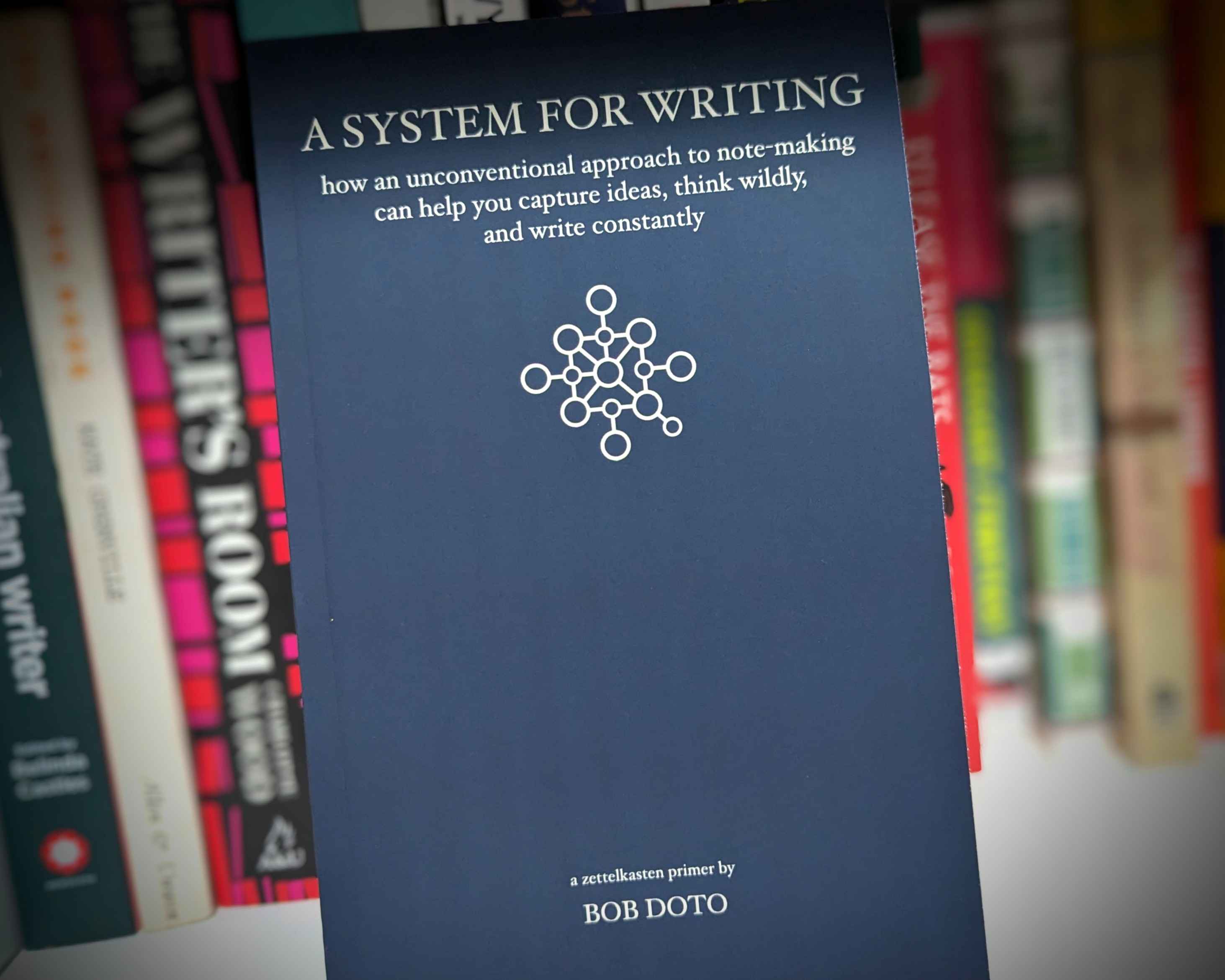 the book cover of A System for Writing by Bob Doto. In the out-of-focus background are book spines in a bookcase