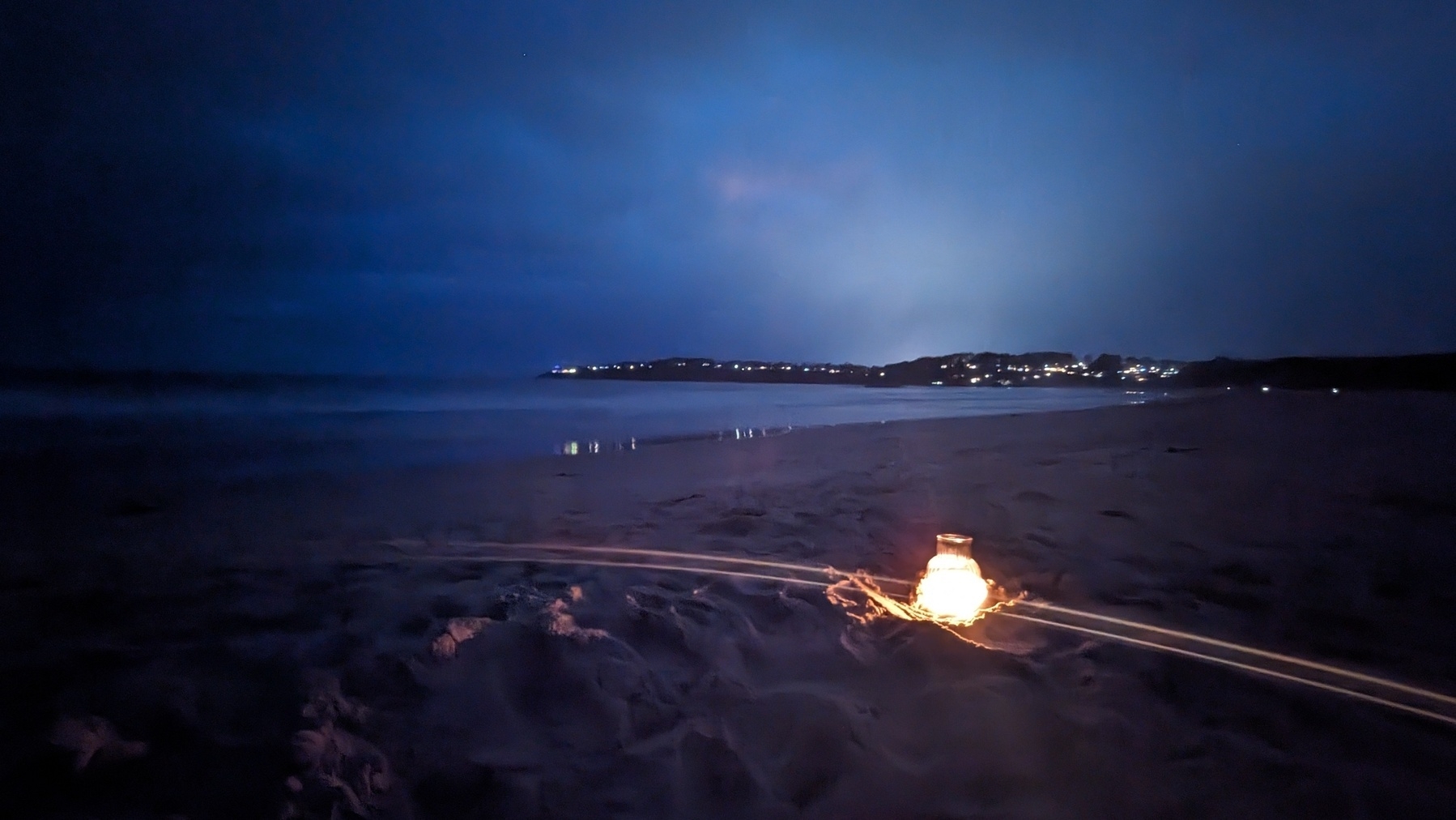 A lantern shines on a beach in the dark. Lights shine on the distant shoreline.