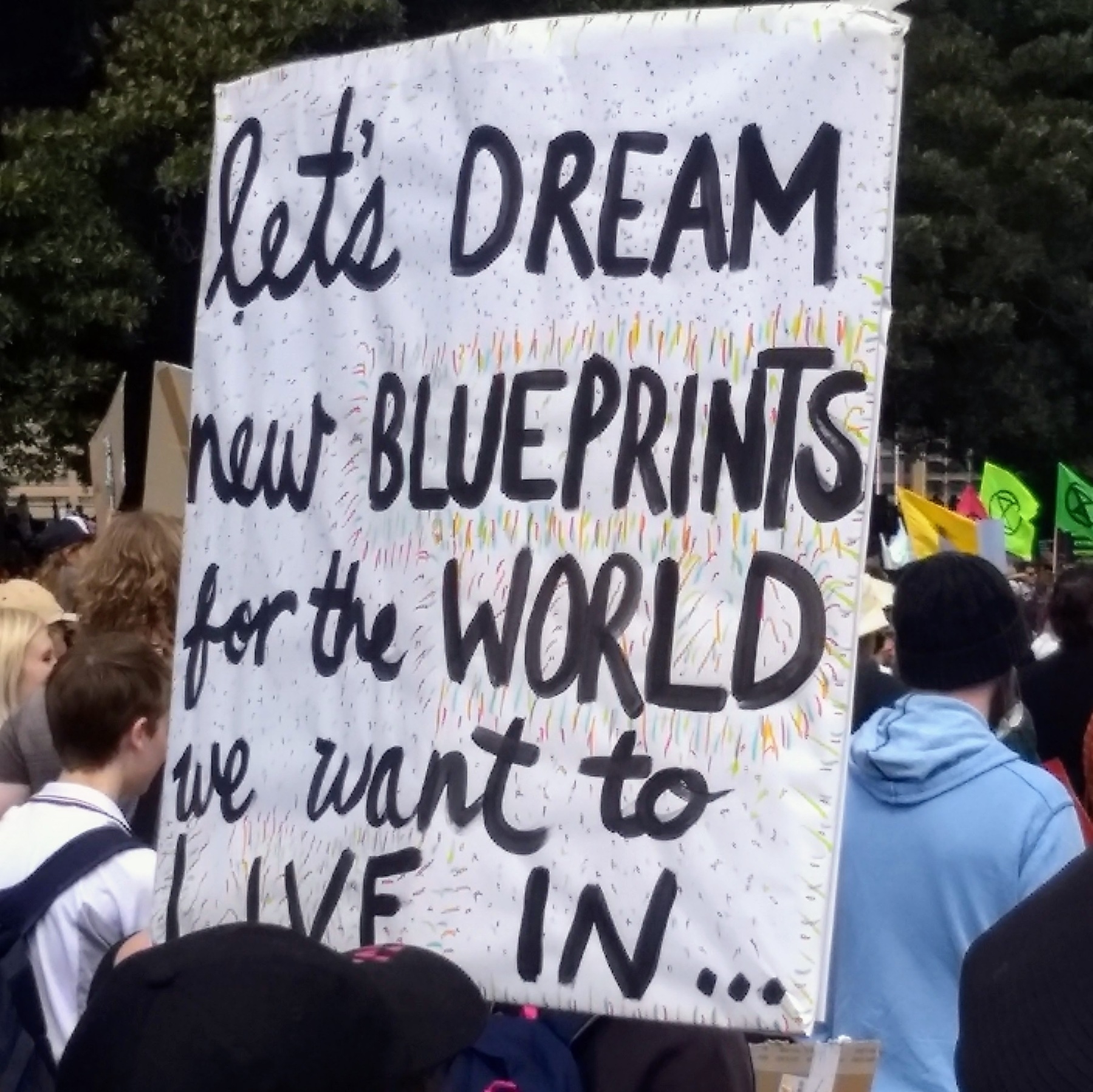 A hand-lettered poster at a rally says Let's dream new blueprints for the world we want to live in...