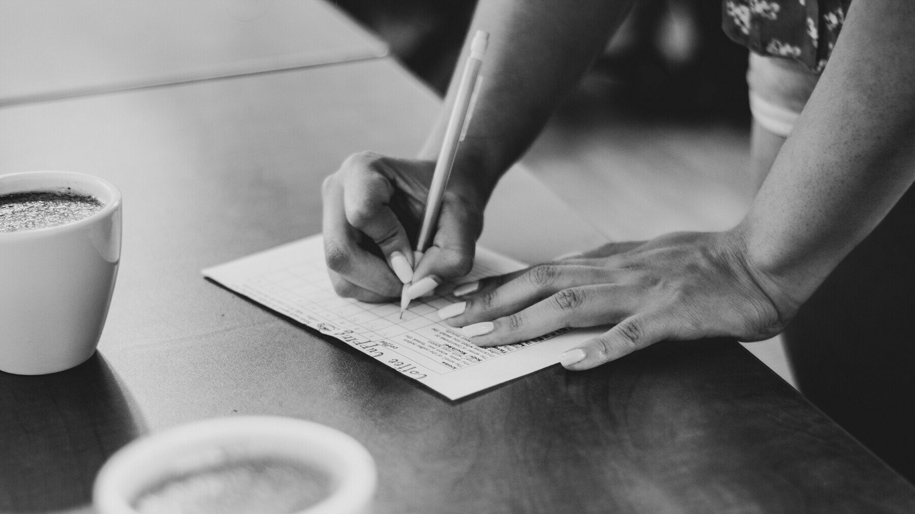 A close-up of someone writing notes with a pen at a table with coffee mugs