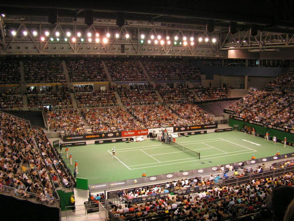 An audience of 10,000 watches a professional tennis match in a well-lit indoor stadium.