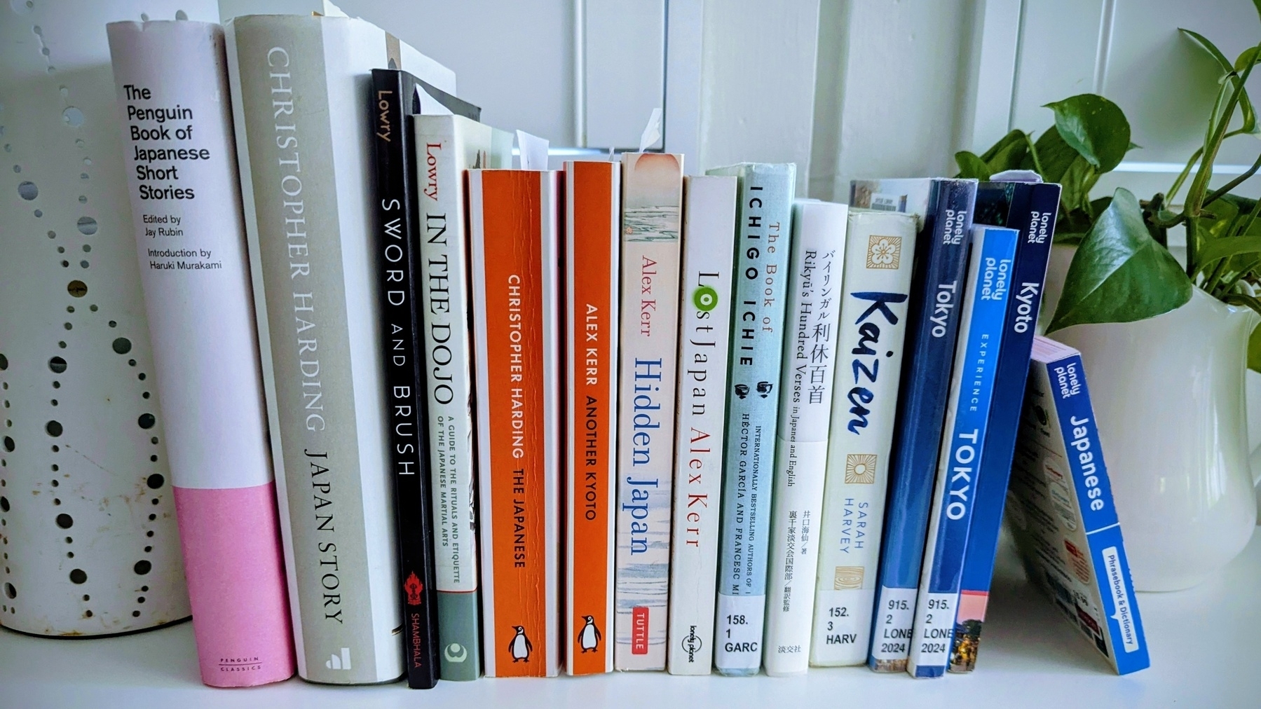 Fifteen books about Japan, spines upright on a white surface next to a leafy house plant.