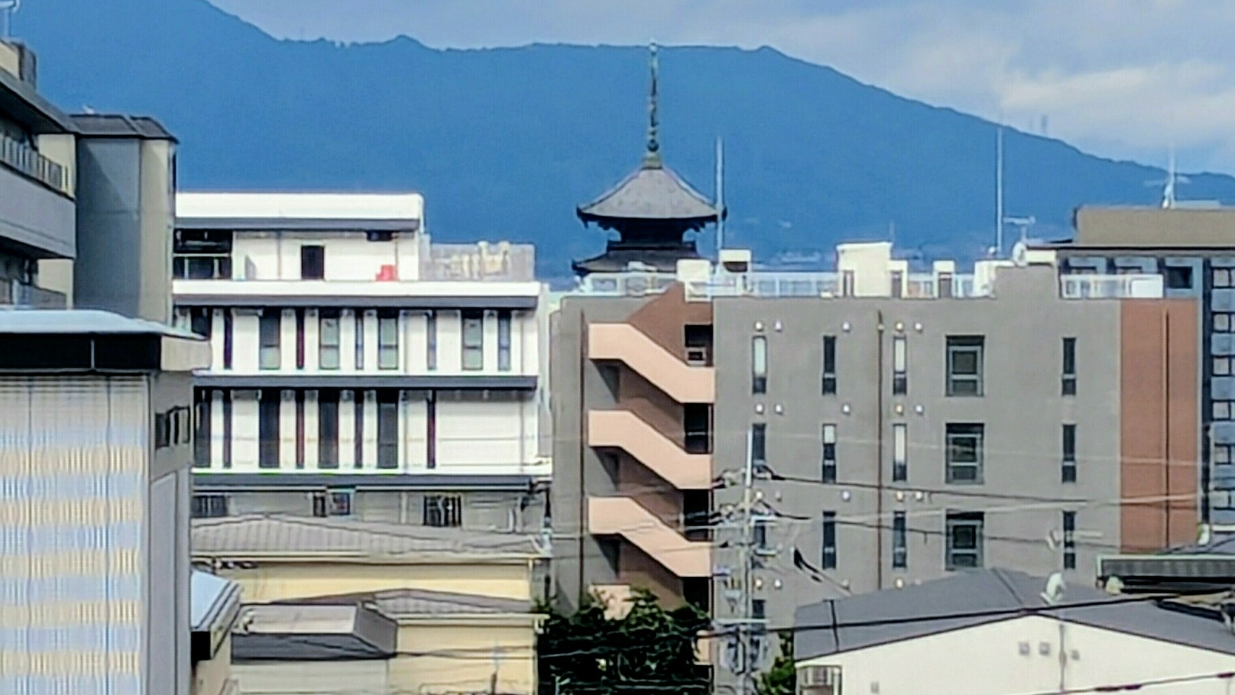 A Kyoto cityscape features modern buildings with a traditional pagoda visible in the distance, set against a backdrop of mountains.