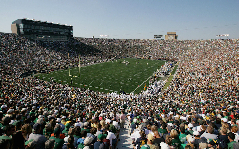 A large crowd of 75,000 fills the stadium, watching a football game in progress on a sunny day.