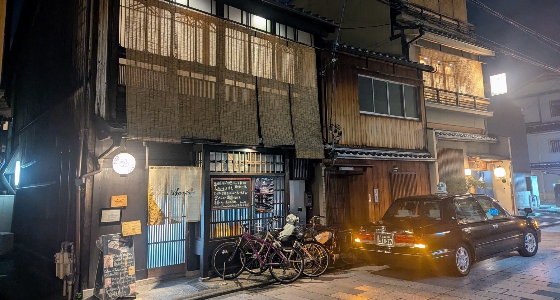 A nighttime street scene in Japan features traditional wooden buildings, a parked taxi, and bicycles against the storefront.