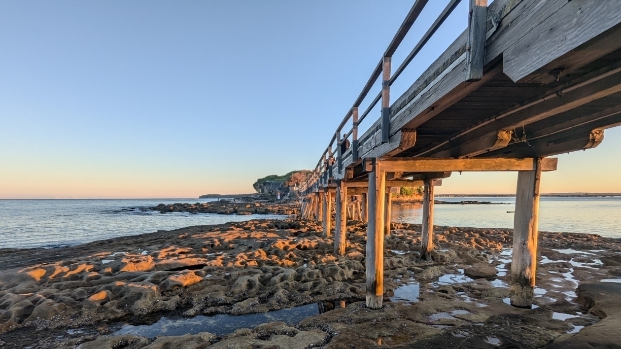 Joining Bare Island to the mainland at La Perouse, Sydney, a wooden bridge extends over a rocky shoreline beside a calm ocean at sunset.