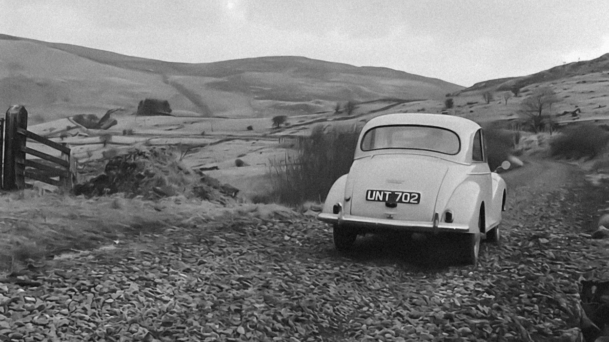 A vintage car is driving along a former railway line near Trawsfynydd, Wales, through a rural landscape with rolling hills in the background.