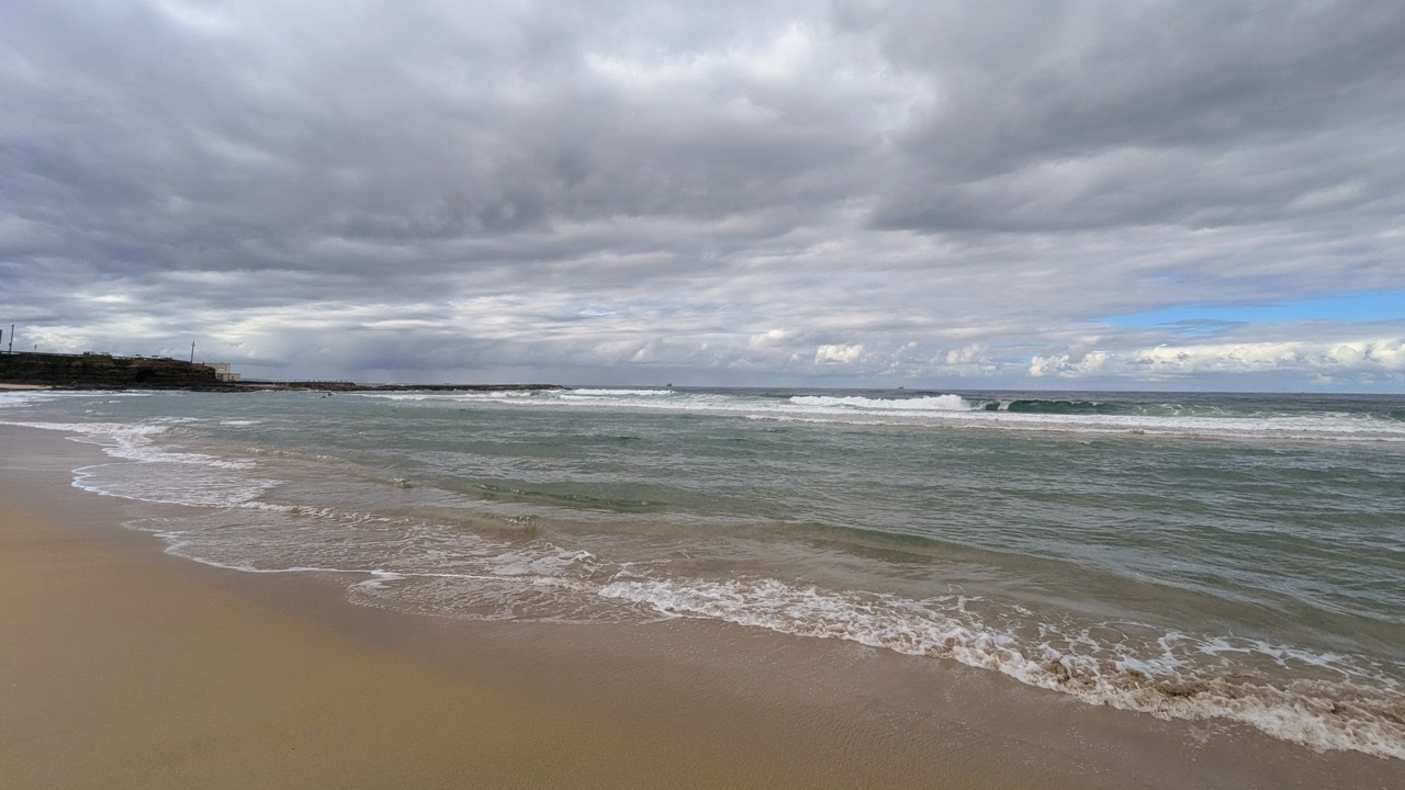 Gentle ocean waves make it to shore on a sandy beach under a cloudy sky.