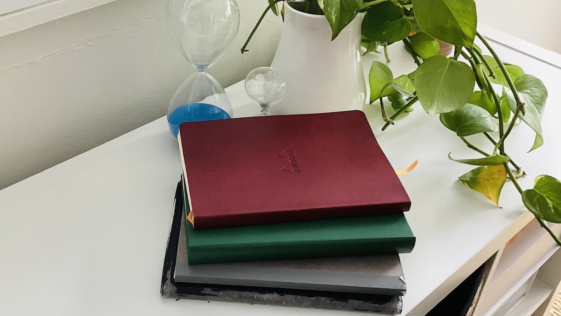 A stack of four notebooks is placed on a white surface next to a potted plant and an hourglass.
