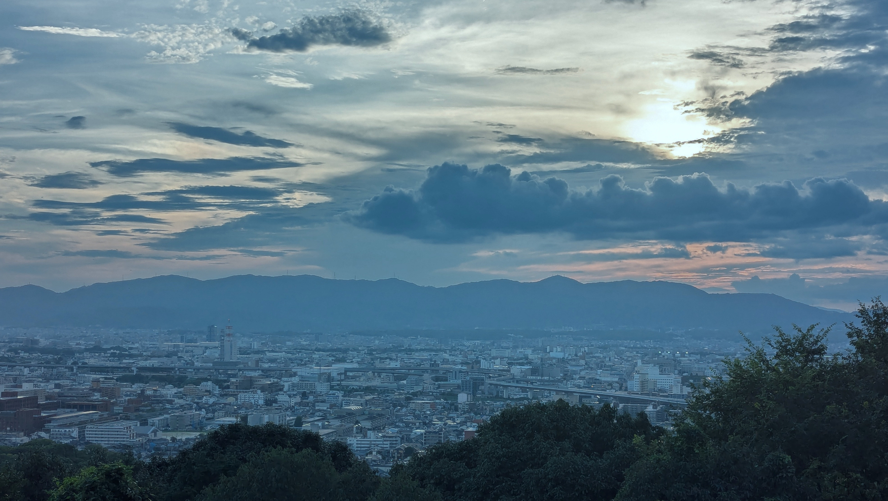 A Kyoto cityscape stretches below a dramatic, cloud-filled sky at sunset, with mountains silhouetted in the background.