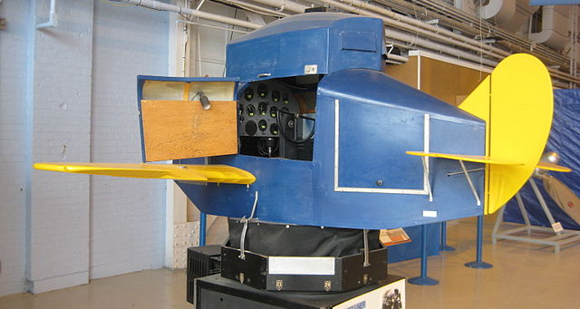 A vintage Link Trainer flight simulator with a blue and yellow exterior is displayed in a Canadian museum, showing an open side panel revealing its controls.