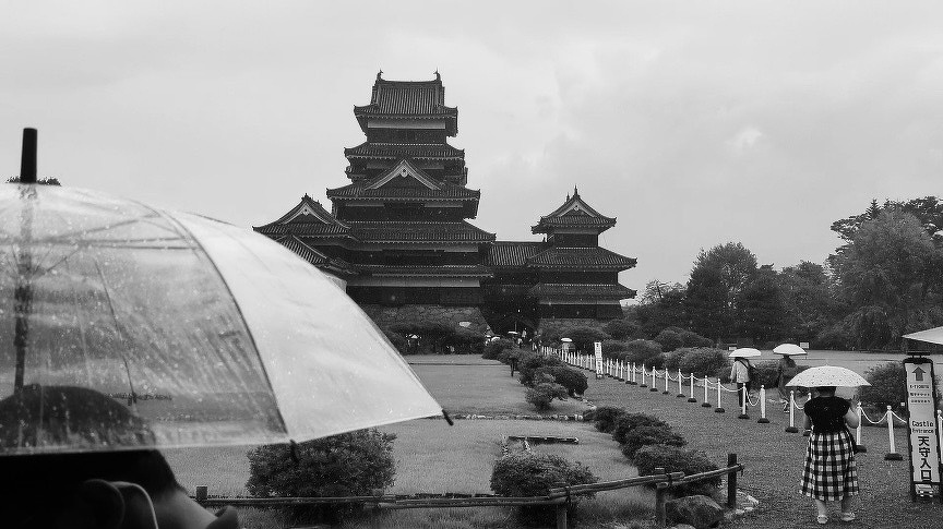 The traditional Japanese castle at Matsumoto stands in the background as people with umbrellas walk through a garden in the foreground on a rainy day.