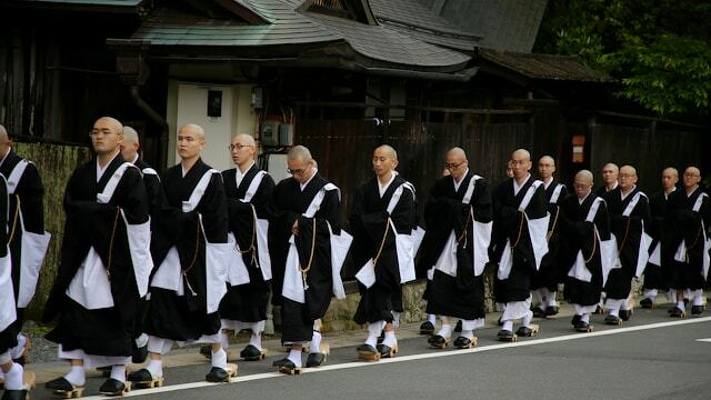 A group of monks dressed in traditional robes and sandals walk in a solemn procession along a street.