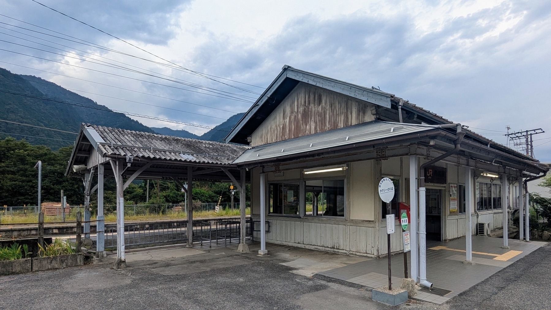 A small, rustic Japanese train station building stands against a backdrop of mountains under a cloudy sky.