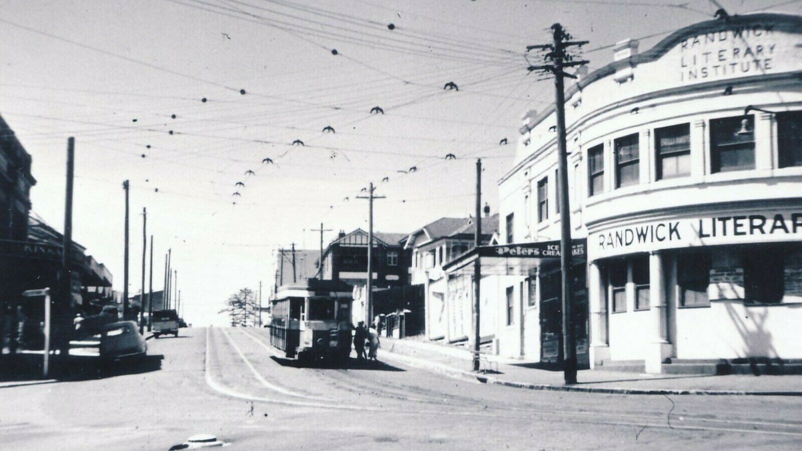 A historic black-and-white street scene features a tram on tracks beside the Randwick Literary Institute building, surrounded by power lines and nearby pedestrians.