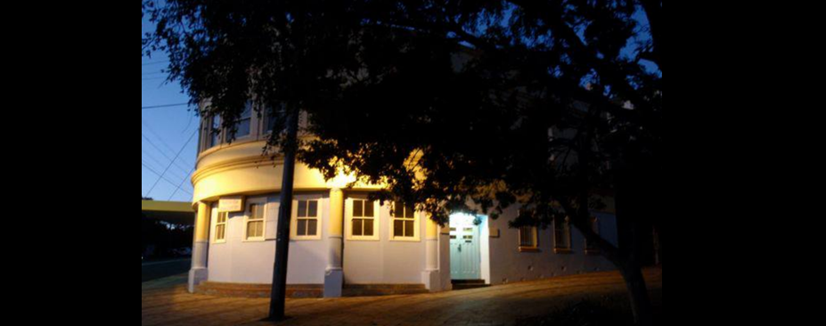 A dimly lit Randwick Literary Institute building is partially obscured by tree shadows under evening light.