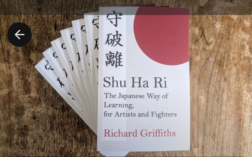 A stack of books titled Shu Ha Ri: The Japanese Way of Learning, for Artists and Fighters by Richard Griffiths is displayed on a wooden surface.